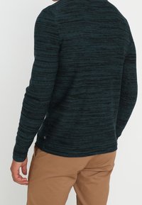 Mörkgrön mönstrad tröja med en texturerad, ribbad design; långa ärmar och rundad halsringning; bärs med khakifärgade byxor.