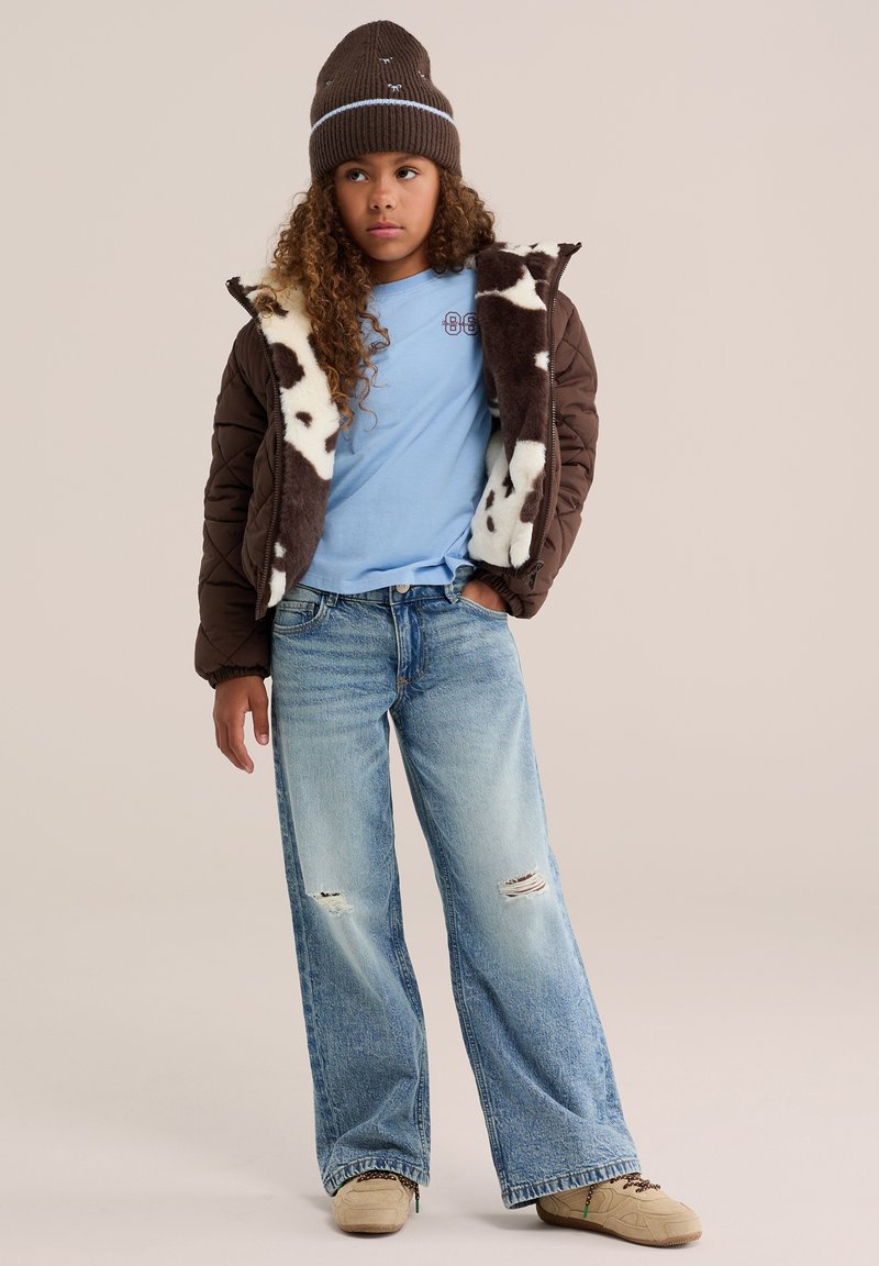 Jeune personne portant un bonnet marron, une veste matelassée marron avec des taches blanches, un t-shirt bleu, un jean large déchiré et des baskets beige, debout avec une main dans la poche.