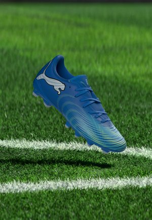 Une chaussure de football bleue Puma flotte au-dessus de l’herbe, à côté d’une ligne blanche du terrain.