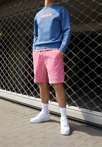 Felpa blu con logo "LEVI'S", pantaloni corti rosa e sneakers bianchi abbinati a calzini alla caviglia. Posizionata di fronte a uno sfondo in metallo testurizzato.