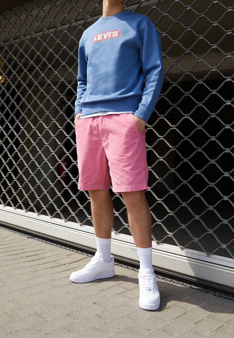 Felpa blu con logo "LEVI'S", pantaloni corti rosa e sneakers bianchi abbinati a calzini alla caviglia. Posizionata di fronte a uno sfondo in metallo testurizzato.