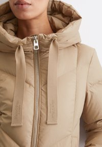 Beige puffer jacket with a high neck, hood, and silver zipper. Features logo accents and diagonal quilted pattern on soft fabric.