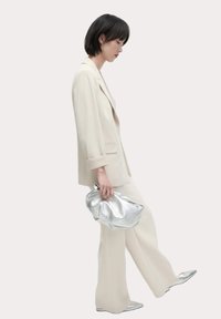 Abito su misura beige chiaro con pantaloni a gamba larga, abbinato a una pochette metallica argentata e scarpe a punta argentate. Texture liscia e design moderno.