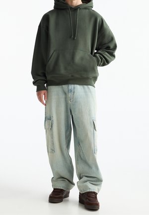 Ciemnozielona bluza z kapturem i przednią kieszenią, zestawiona z jasnoniebieskimi spodniami typu cargo z bocznymi kieszeniami oraz brązowymi tenisówkami.