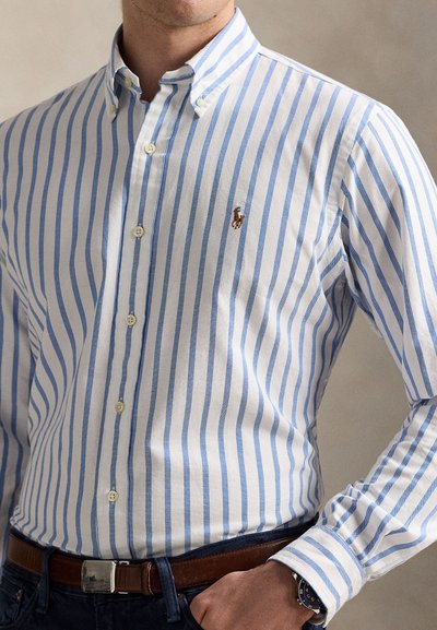 Bela in svetlo modra srajca z gumbi in ovratnikom, z majhno vstavljenim logotipom in nežno teksturo. Nošena s rjavim pasom.