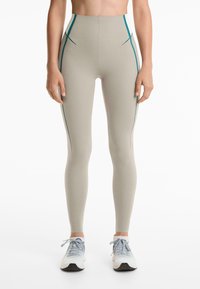 Vrouw in hoge beige legging met teal zijstrepen en witte sportschoenen, staande tegen een witte achtergrond.