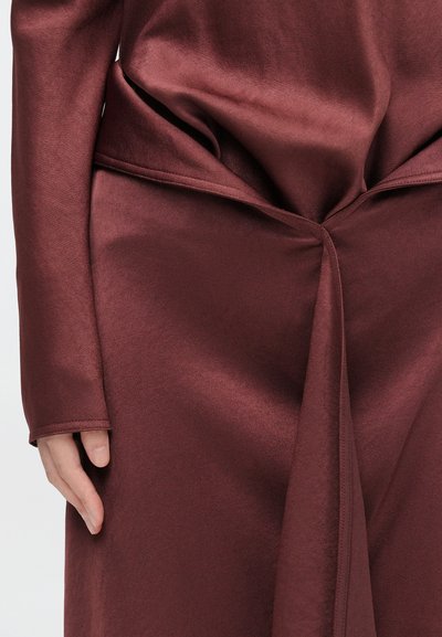 Peça de vestuário bordô com uma textura lisa, semelhante a cetim; apresenta pregas drapeadas e um design sem emendas; mão parcialmente visível ao lado.