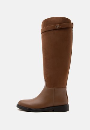 Botte d'équitation marron montant jusqu'au genou avec pied en cuir lisse, dessus en suède, talon bas noir et sangle décorative avec détail de logo métallique.