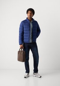 Blauer Daunenjacke mit hohem Kragen, darunter ein hellblaues Hemd, dazu dunkle Jeans und eine braune Ledertasche. Trägt silberne Sneakers.