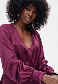 Bordowa sukienka z długim rękawem i dekoltem w serek, o jedwabistej fakturze, ze ściąganymi ramionami i obszernymi rękawami, podkreślona delikatnymi elastycznymi mankietami.