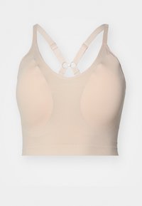 Beige, sømløs BH med bredt belte, formede kopper og kryssende stropper med en metallring som accent bak. Mykt, stretchy stoff.