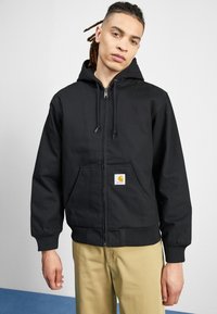 Black zip-up hoodie made of durable fabric, featuring a front pocket and ribbed cuffs. Notable Carhartt logo on the front.