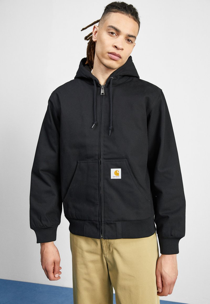 Black zip-up hoodie made of durable fabric, featuring a front pocket and ribbed cuffs. Notable Carhartt logo on the front.