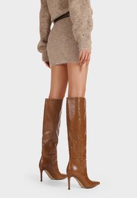 Stivali alti marroni con effetto coccodrillo, con punte affusolate e design aderente fino al ginocchio, abbinati a una mini gonna di maglia beige.