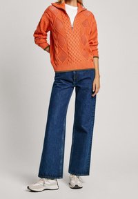 Jersey de punto naranja con cuello con cremallera, que presenta un patrón de trenzas. Combinado con vaqueros de pierna ancha azul y zapatillas beige.