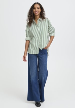 Camicia a righe verdi e bianche con maniche arrotolate, abbinata a pantaloni in denim blu scuro a gamba larga e stivali neri.