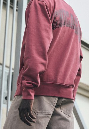 Persoon die een verweerde rode trui draagt met de tekst "CHICAGO" op de achterkant en lichtbeige broek, staand bij een gebouw met ramen.