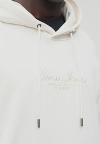 Sudadera blanca con capucha que presenta un logotipo bordado de "Guess Jeans", cordones con puntas de metal y una textura suave.