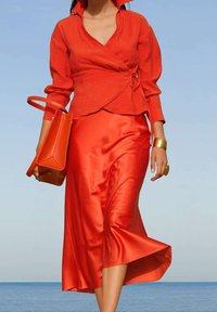 Femme marchant près de l'eau portant un chemisier portefeuille rouge et une jupe en satin, portant un sac à main rouge, avec des accessoires bracelet et bague en or.