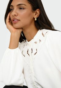 Kvinde med mørkt hår iført guld hoops og en hvid bluse med blonde detaljer, som hviler hagen på sin hånd.