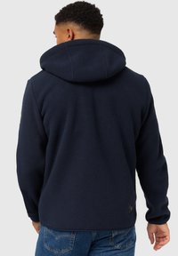 Tumesine fleece-kapuga kraeaga sviiter, millel on tekstuuriga viimistlus, reguleeritav kapuuts ja elastsed mansetid. Seljal allosas on väike brändi silt.