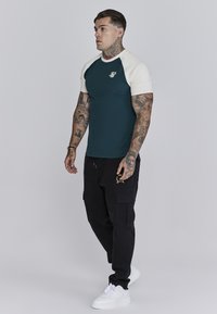 Korte mouwen, strak aangesloten teal t-shirt met crèmekleurige raglanmouwen en een logo. Gepaard met zwarte cargo pants en witte sneakers.