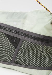 Green nylon bag with a black mesh pocket, features a gray zipper and colorful braided strap. Includes logo detail near the zipper.