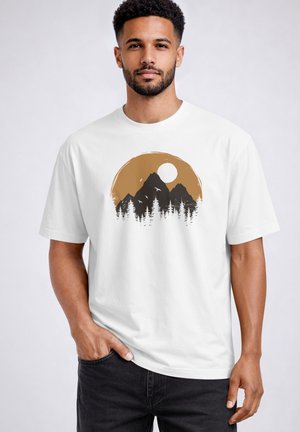 Mann trägt weißes T-Shirt mit Grafik von Bergen, Bäumen, Vögeln und Sonne in Braun- und Schwarzfarben, steht vor einfachem hellem Hintergrund.