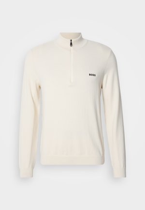 Jasnobeżowy sweter z dzianiny z wysokim kołnierzem i półsupełkiem, z małym czarnym logo na lewej piersi i ściągaczami przy rękawach.