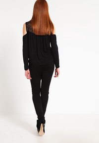 Svart off-shoulder topp med spetsdetaljer, i kombination med svarta skinny jeans och svarta klackstövlar, sedd bakifrån.