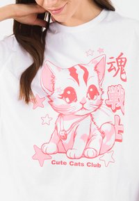 Balta T-kreklu ar rozā krāsā izdrukātu karikatūras kaķi, apbrīnotu ar zvaigznēm un japāņu rakstiem, kas rāda rotaļīgu dizainu ar zvana aksesuāru.
