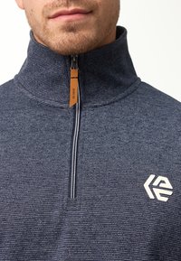 Marineblå zip-up pullover i tekstureret stof med hvide striber, der har en høj krave og logo broderet på venstre bryst. Lynlåstrækkeren har en læderdetalje.