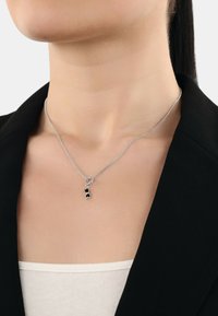 Collana d'argento con un pendente a forma di anello, caratterizzata da un piccolo ciondolo nero. Indossata attorno al collo sopra una maglietta beige.