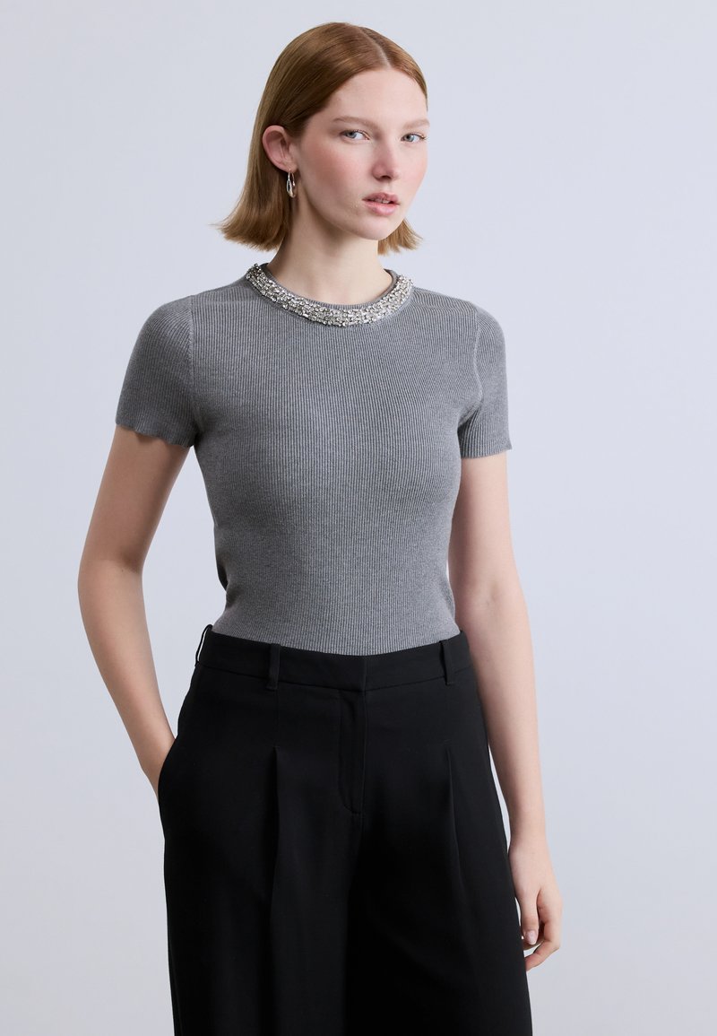 Γκρι ριμπ κοντομάνικη μπλούζα με κυματιστό τελείωμα και διακοσμημένο ντεκολτέ, συνδυασμένη με μαύρο ψηλόμεσο παντελόνι με πιέτες.