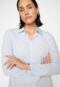 Világoskék csíkos ing hosszú ujjal, klasszikus gallérral és fodros mandzsettával. Simább, könnyű anyagból készült.