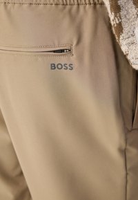 Beige byxtyg med en slät textur, som har en ficka med dragkedja och en grå "BOSS"-logotyp tryckt i närheten.