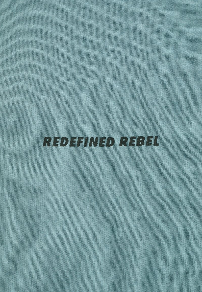 Helle blaue Baumwollstoffe mit dem Text "REDEFINED REBEL" in fetter schwarzer Schrift, zentral auf der Oberfläche des Materials platziert.