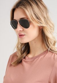 Ochelari de soare tip aviator cu lentile întunecate, cadru subțire din argint și detalii subtile. Purtați cu un top roz deschis, evidențiind textura netedă a materialului.