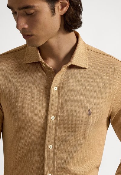 Koszula zapinana na guziki z wzorem jodełki w ciepłym odcieniu brązu. Posiada szpiczasty kołnierzyk oraz guziki dopasowane do tkaniny. Logo na piersi.