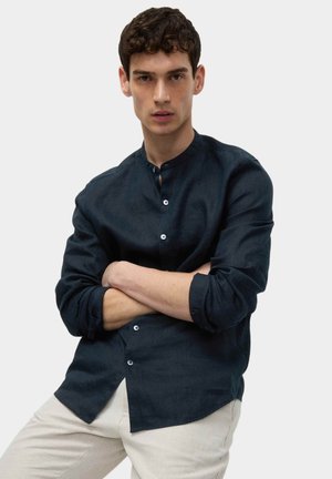 Junger Mann mit verschränkten Armen, der ein dunkelmarineblaues Hemd mit Stehkragen und hellbeige Hosen vor einem schlichten Hintergrund trägt.