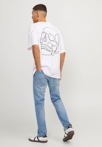 Hvit bomulls T-skjorte med svart linje hodeskallegrafikk på baksiden, kombinert med lyseblå denimjeans og svart-hvite sneakers.