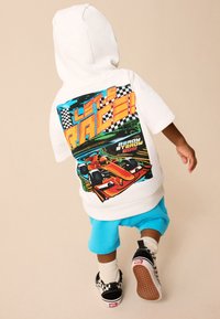 Balta džemperio su gaubtu su spalvotu lenktynių grafiku ant nugaros, derinama su šviesiai mėlynais šortais ir juodais sportbačiais su languotu raštu.