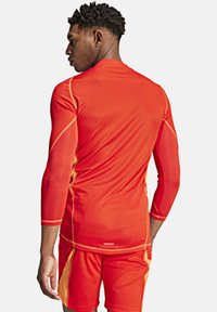 Rode sportieve longsleeve met oranje accenten, textuurstof en aansluitend ontwerp. Draagt met bijpassende shorts die de sportieve snit benadrukken.