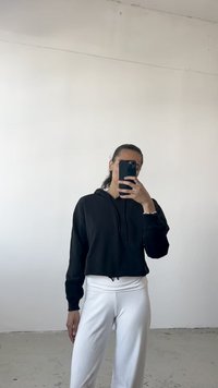 Sweatshirt preto cortado com capuz ajustável, combinado com calças brancas de cintura alta. Textura suave, design descontraído, situado contra um fundo simples.