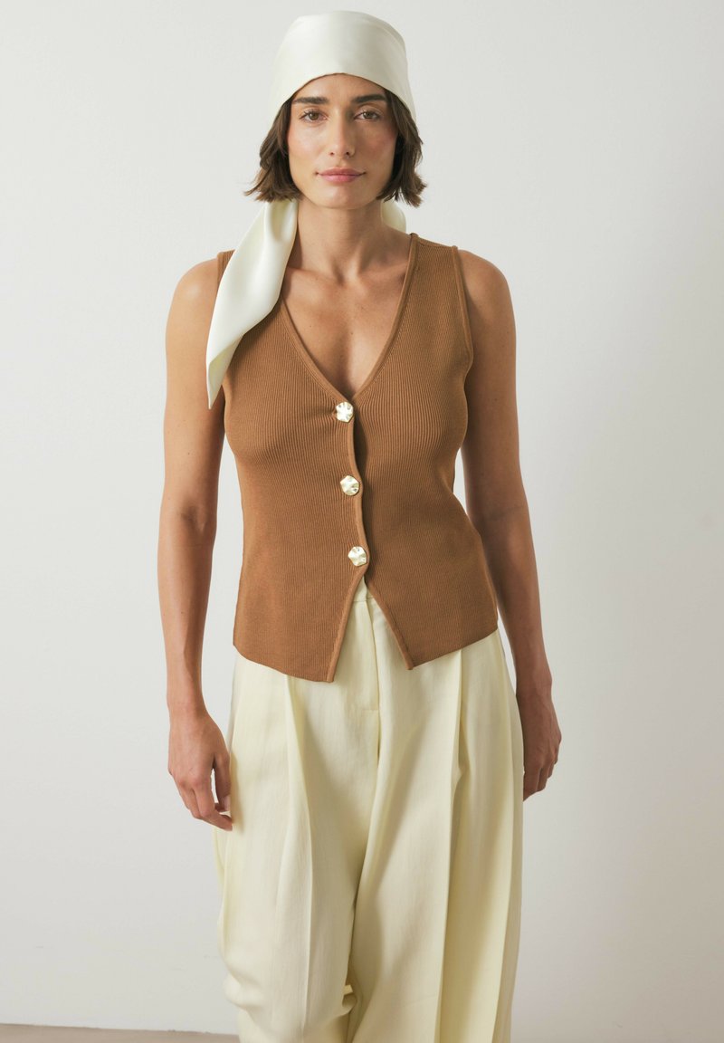 Donna con un velo color crema, top senza maniche marrone con bottoni e pantaloni larghi color crema, in piedi contro uno sfondo chiaro e uniforme.
