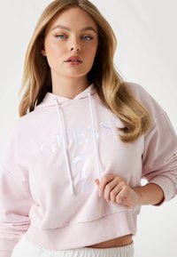 Cropped hoodie rosa chiaro con cordini bianchi, dotato del logo "GUESS" in rilievo e design di punto interrogativo sulla parte anteriore. Tessuto morbido e orlo a coste.