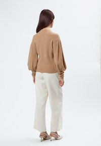 Beige gestrickter Pullover mit Ballonärmeln, kombiniert mit lockeren weißen Hosen und hellen, höheren Schuhen. Einfaches Design, strukturierter Stoff.