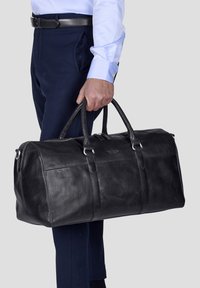 Black leather duffel bag with dual handles, zipper closure, and visible stitched accents, held in a hand, displaying a smooth texture.