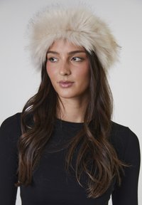 Faux-Fur-Hut in hellem Beige mit einem flauschigen, voluminösen Rand; glatte Stoffbasis; getragen mit einem schwarzen Langarmoberteil; neutraler Hintergrund.
