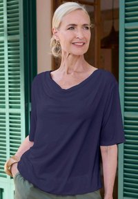 Marineblauwe blouse met een gedrapeerde halslijn en korte mouwen, gemaakt van zachte stof. Draagt met lichtgekleurde broek en een accentarmband.